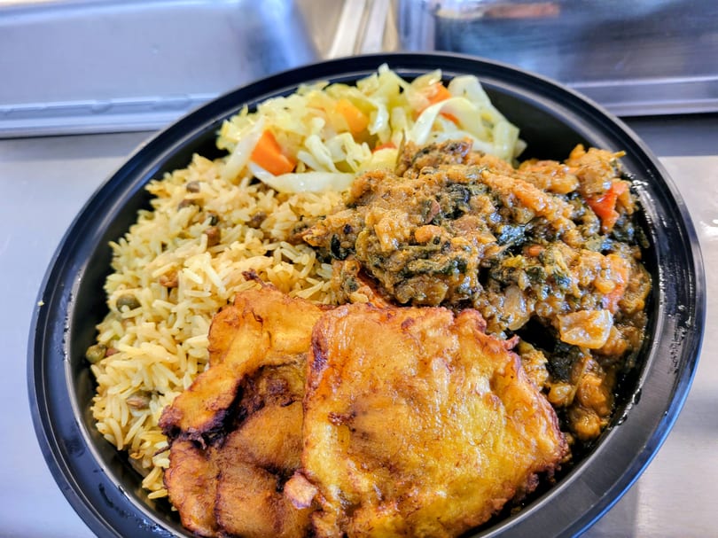 Haitian meal with rice, stewed greens, fried plantains, and cabbage in a black bowl.