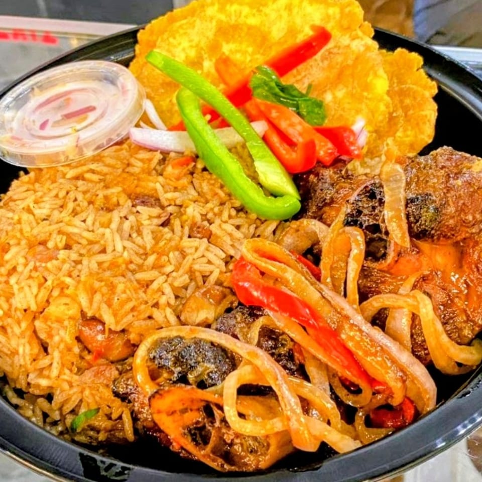 Haitian meal with rice, stewed meat, fried plantains, and pickled vegetables in a black bowl.