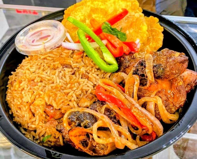 Haitian meal with rice, stewed meat, fried plantains, and pickled vegetables in a black bowl.