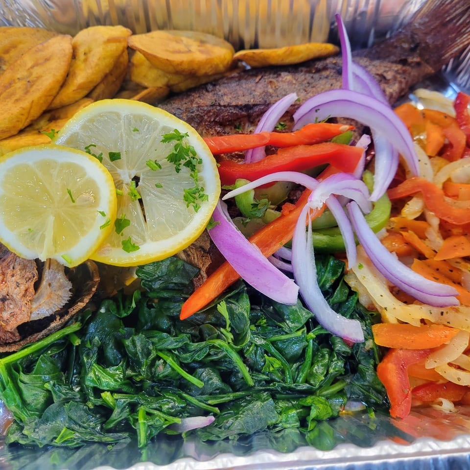 Haitian fried fish with plantains, spinach, and pickled vegetables in a foil container.