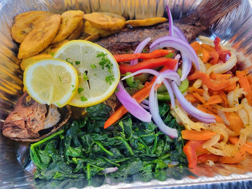 Haitian fried fish with plantains, spinach, and pickled vegetables in a foil container.